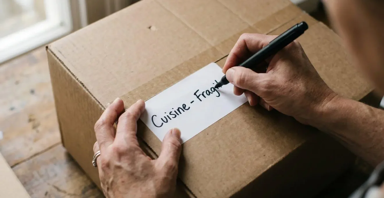 Gros plan sur une main en train d'écrire au marqueur noir sur un carton de déménagement posé sur une table en bois clair